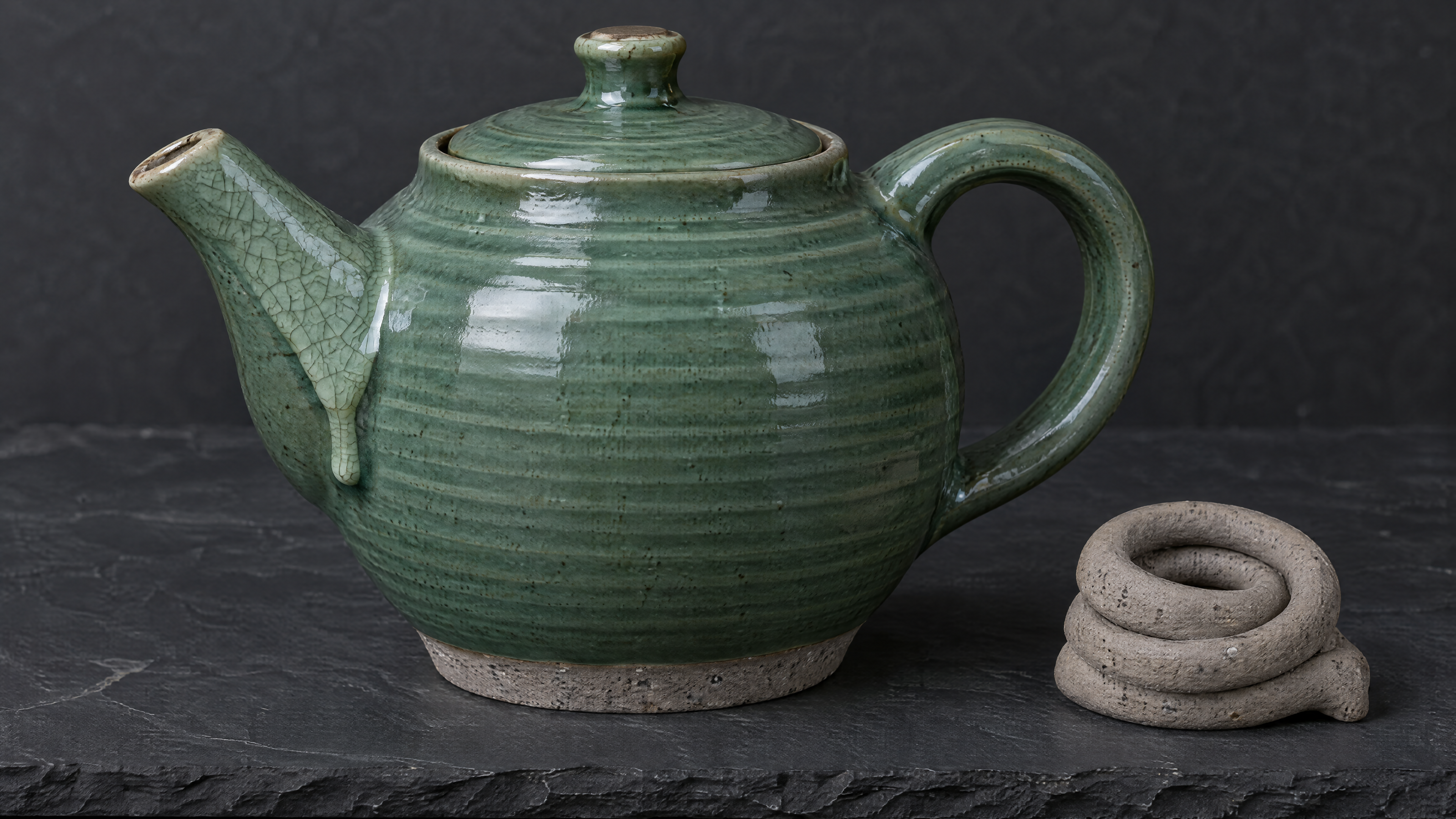 An evenly lit product photograph of a hand-thrown ceramic teapot glazed in deep celadon green, resting on a dark slate surface. Visible spiral throwing rings o…